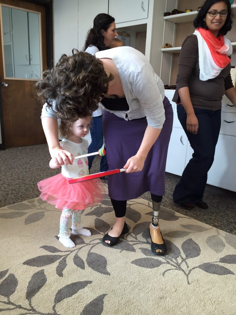 Toddler playing rhythms on lollipop drum with teacher while other students march in a circle to the beat